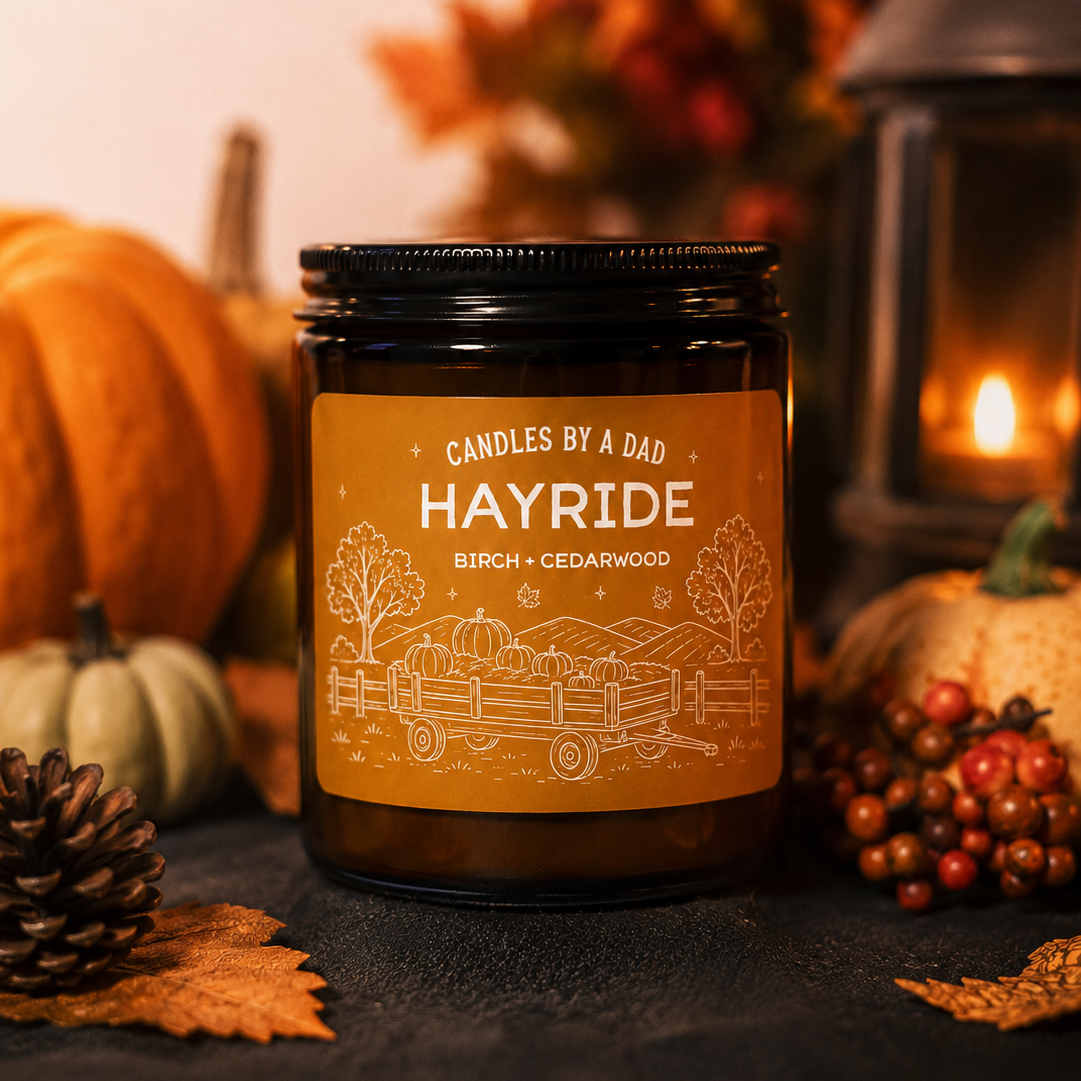 A dark amber glass jar candle with a black lid and a warm orange label reading 'Candles by a Dad Hayride Birch + Cedarwood,' styled with pumpkins, autumn leaves, pinecones, and a lantern in a cozy fall setting.
