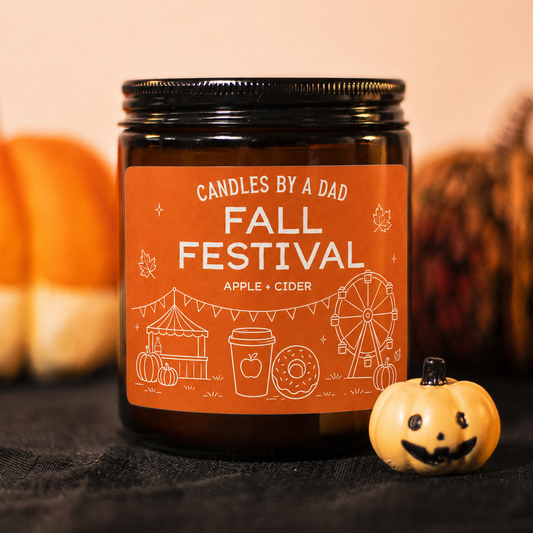 An amber glass jar candle with a black lid and an orange label reading 'Candles by a Dad Fall Festival Apple Cider,' styled on a dark fabric surface with pumpkins and autumn decor blurred in the background.