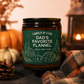A jar candle labeled 'Dad's Favorite Flannel' by Candles by a Dad, styled on a dark fabric surface with autumn props including pumpkins, a pinecone, a lit lantern, and plaid fabric.