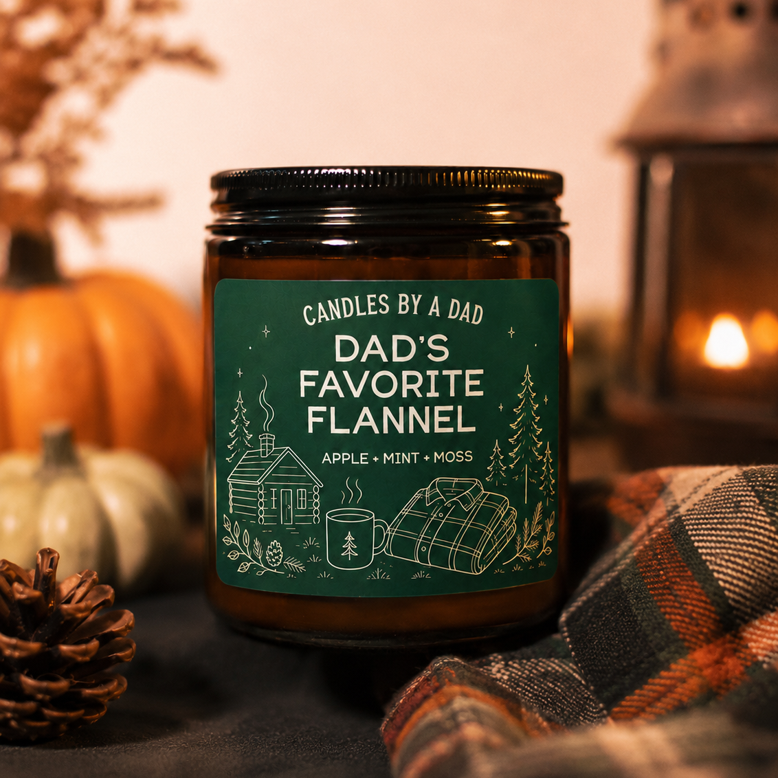A jar candle labeled 'Dad's Favorite Flannel' by Candles by a Dad, styled on a dark fabric surface with autumn props including pumpkins, a pinecone, a lit lantern, and plaid fabric.