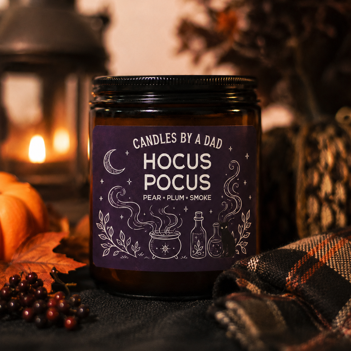 A dark amber glass jar candle with a black lid and a dark purple label reading 'Candles by a Dad Hocus Pocus' with white witch illustrations. The candle is styled on a dark fabric surface with autumn props including a lantern, pumpkins, leaves, and a plaid blanket in a warm, cozy setting.